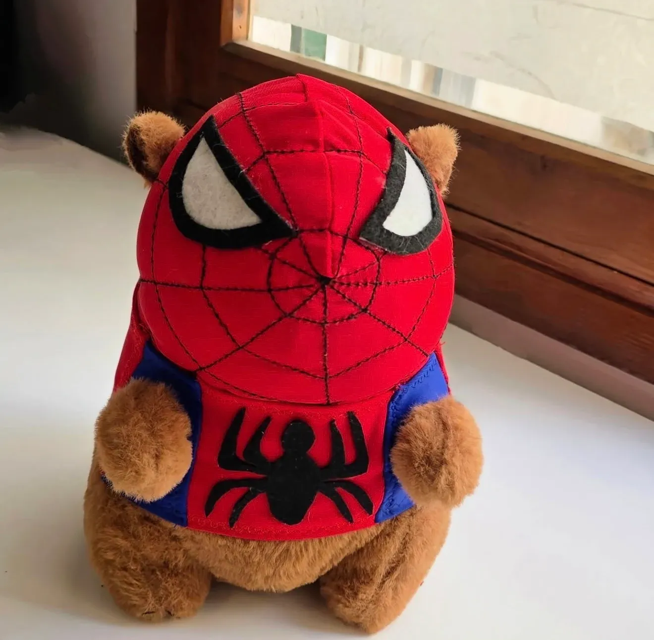 Capibara sentado con traje rojo y azul, emblema de araña negra y ventana de madera al fondo