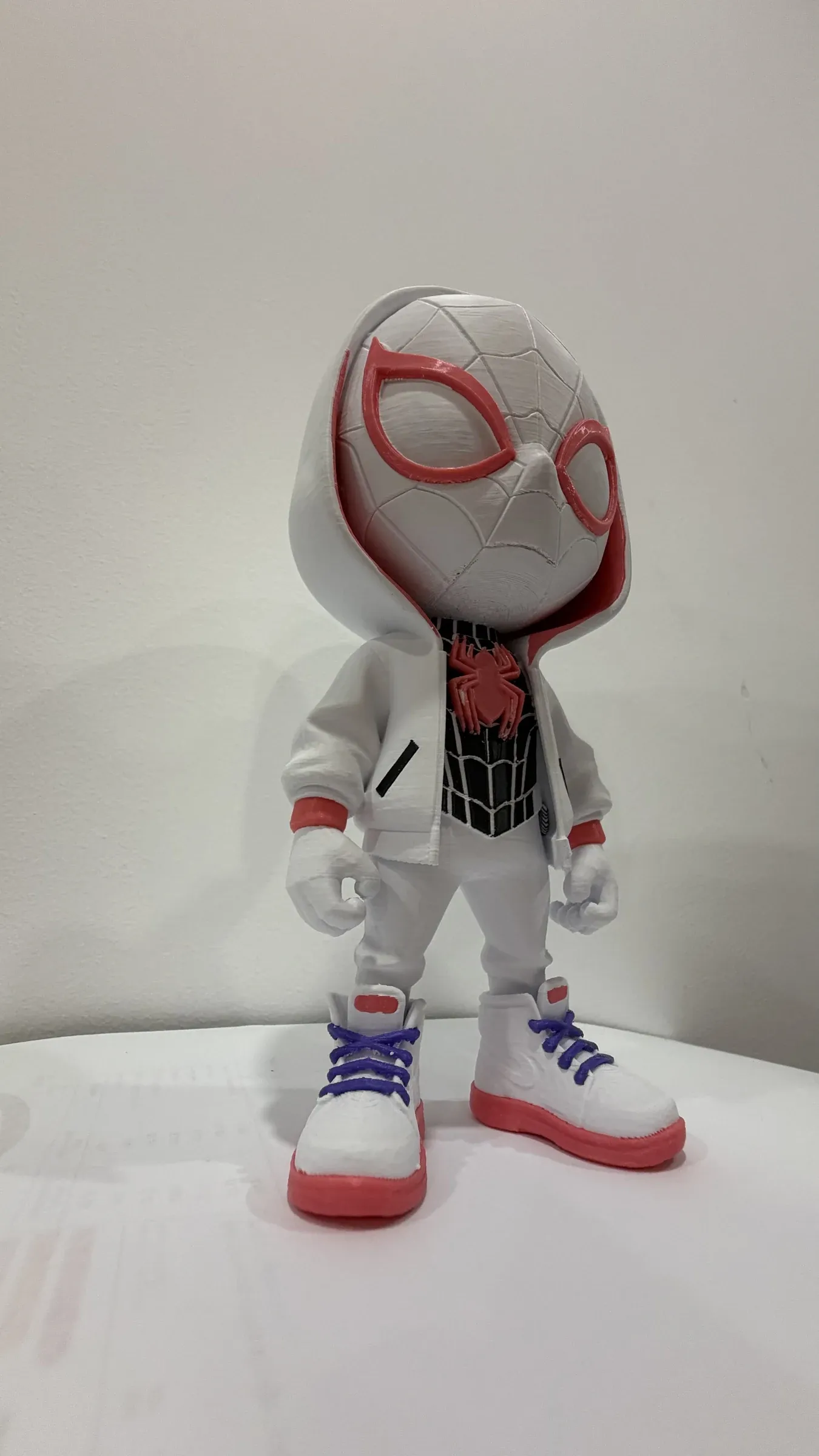 Figura chibi blanca con capucha, cabeza grande y emblema de araña rojo y negro en el pecho.