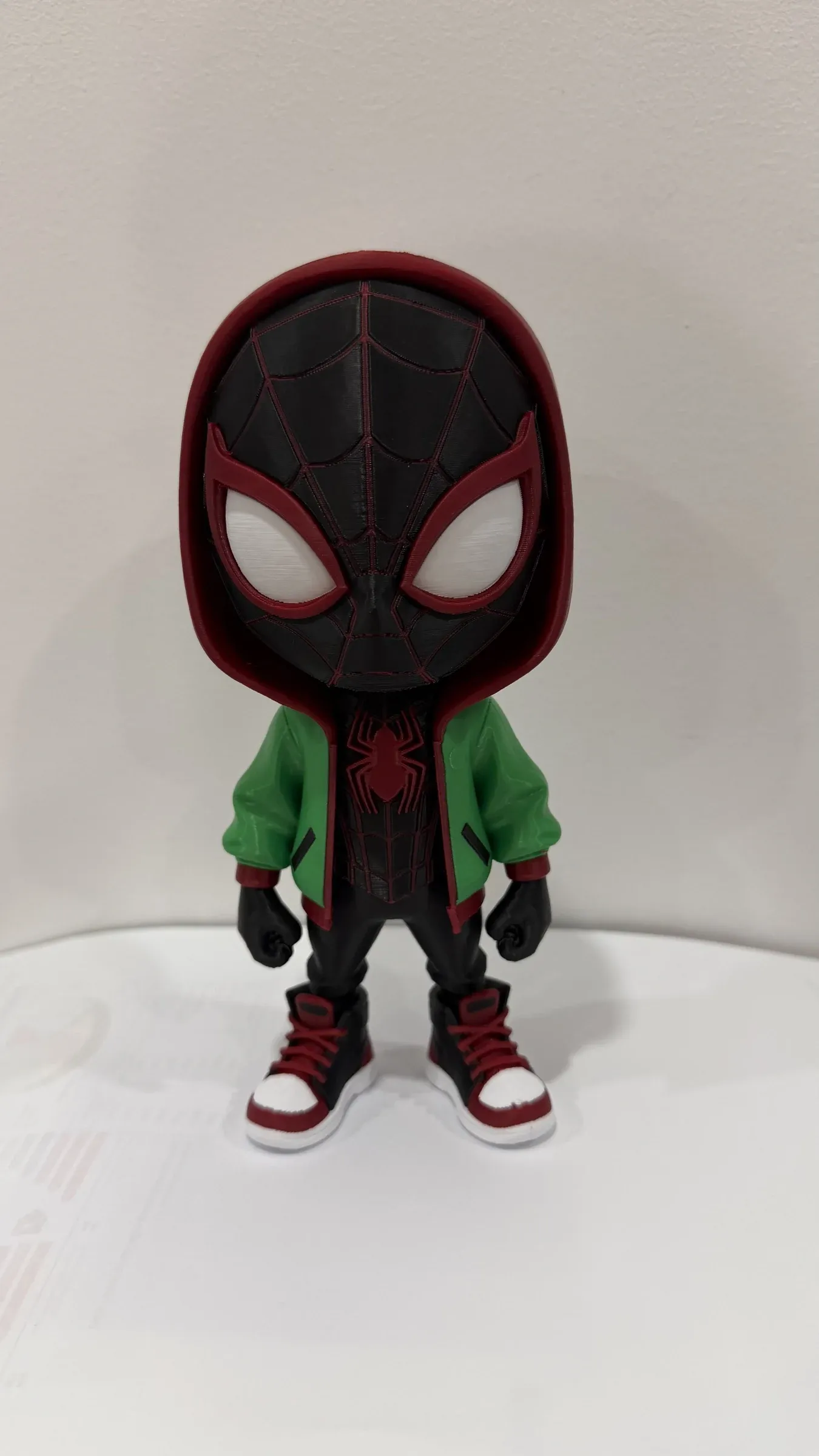 Figura chibi de pie con máscara negra con líneas rojas, ojos blancos grandes y chaqueta verde.