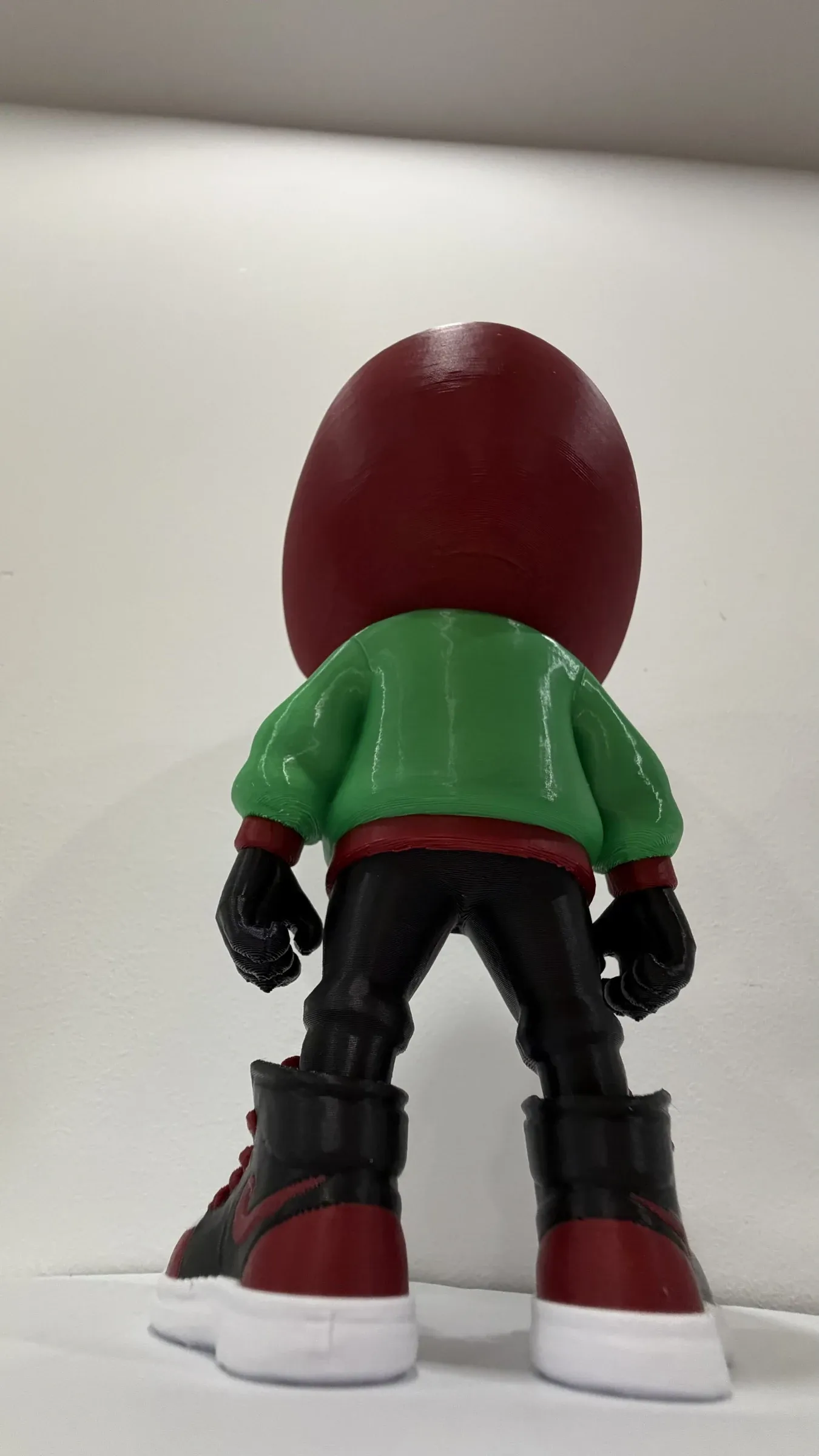 Vista de espalda de la figura, con cabeza grande, chaqueta verde y zapatillas rojas y negras.