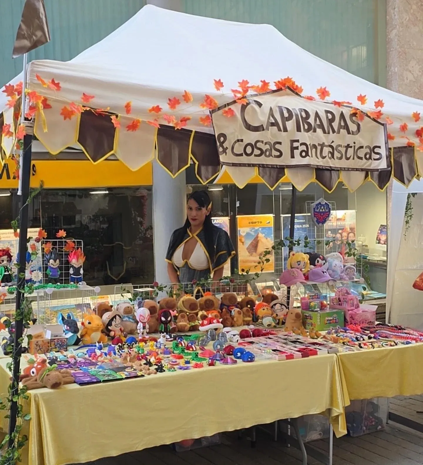 Chigüibara stall with plush toys, 3D figures and geek accessories at a fair, decorated with autumn leaves and a sign that…
