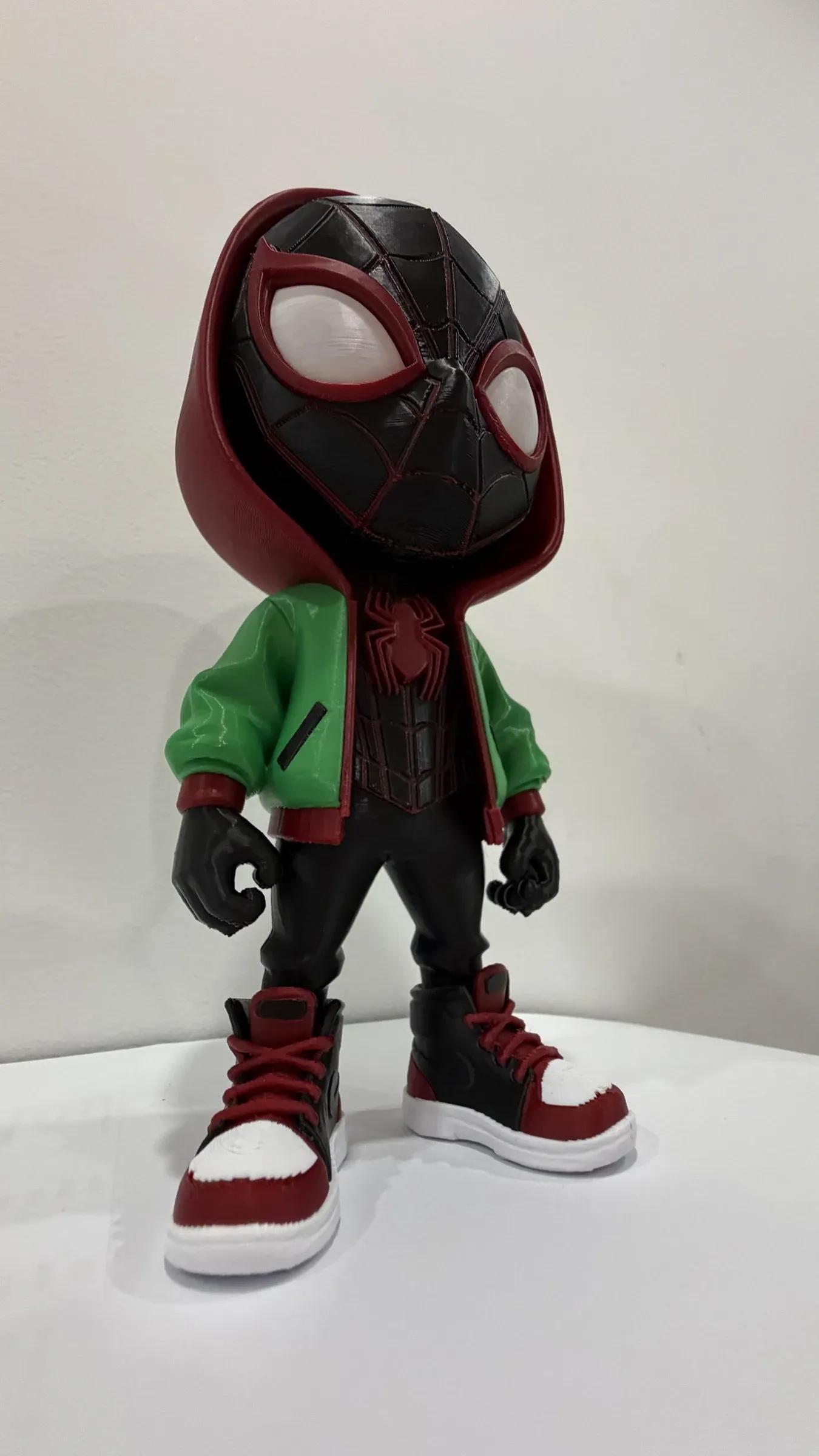 Figura chibi de pie con capucha roja y negra, chaqueta verde y zapatillas rojas y blancas.