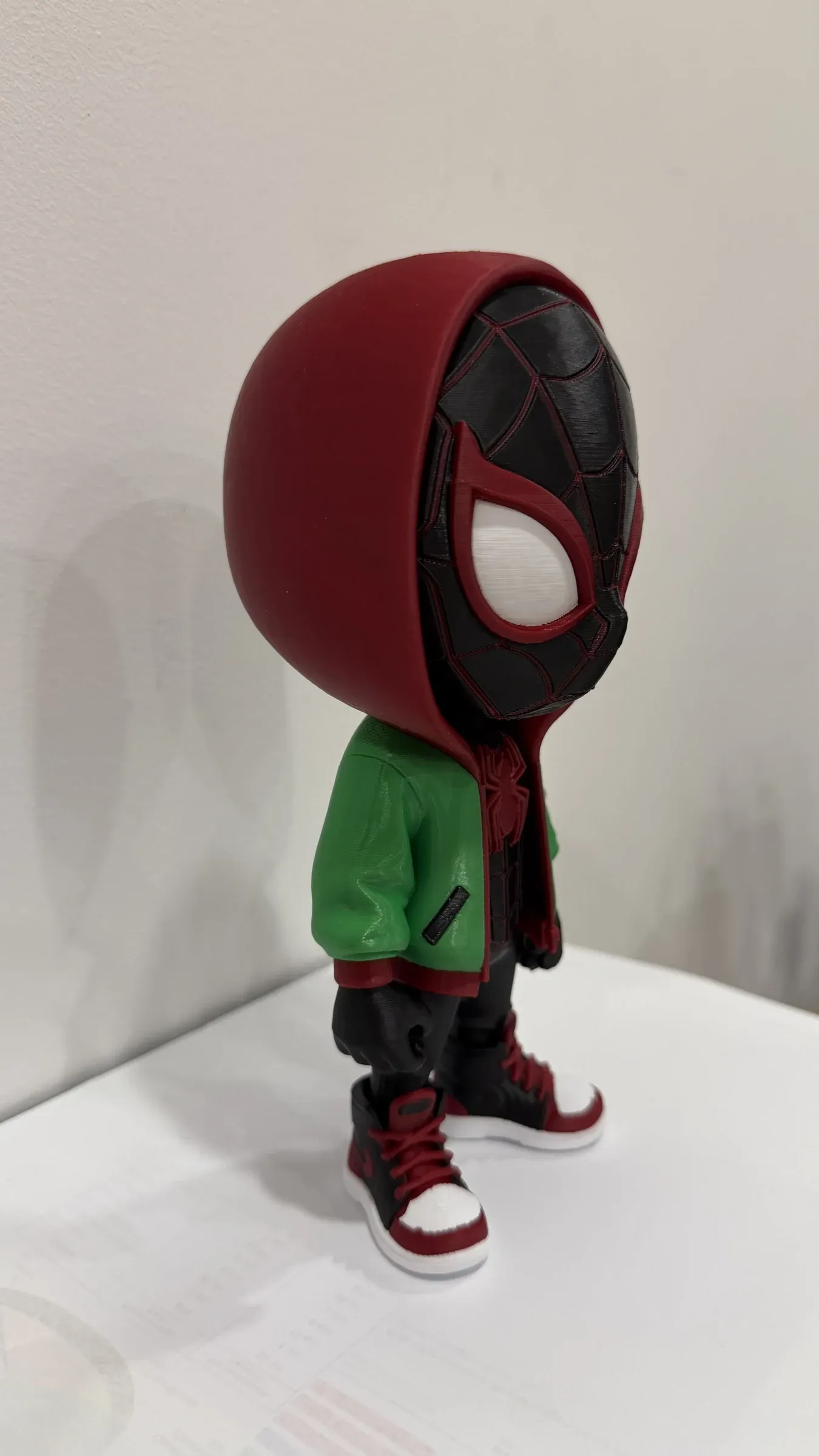 Vista lateral de la figura con capucha roja, chaqueta verde, pantalón negro y zapatillas altas.